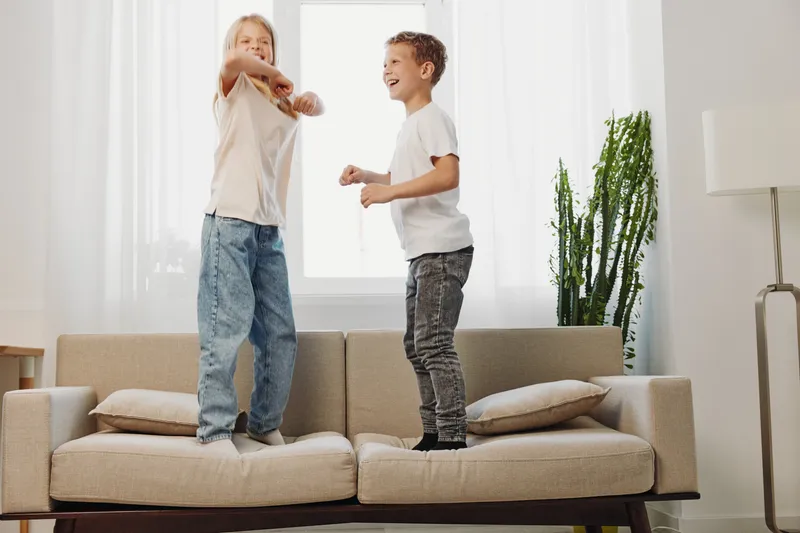 Two active children jump joyfully on a beige sofa in a bright living room with natural light. The scene captures playful energy, childhood fun, and carefree indoor activity.