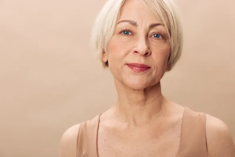 Portrait of mature woman with short blonde hair and natural makeup, wearing beige top against neutral background. Concept for aging beauty, confidence, and simplicity.