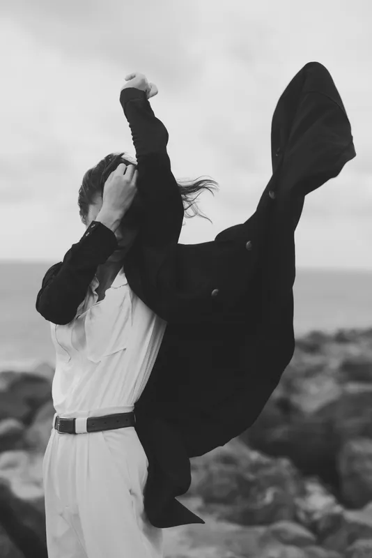 Cape flies, dress swirls in wind along the ocean shore, capturing bold motion and dance with a dramatic silhouette and windblown mood, expressive energy and timeless monochrome style