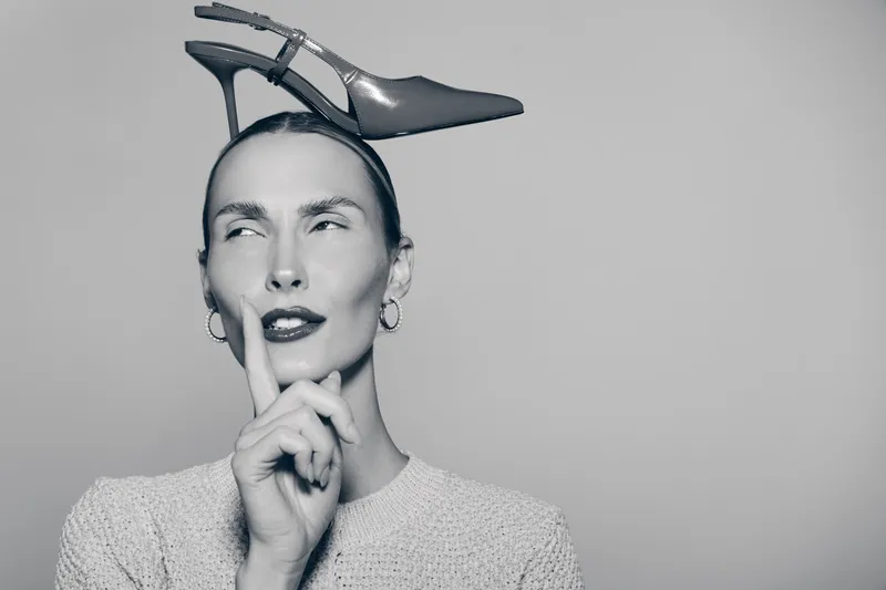 Stylish Caucasian woman with light skin and slicked hair balances a high heel on her head with finger near lips in thoughtful pose. Concept of fashion and creativity.