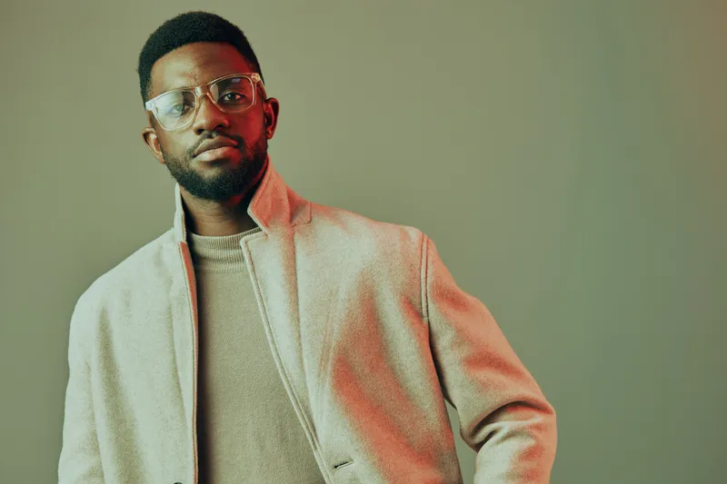 Stylish young African man wearing a beige coat and glasses, posing confidently against a muted gray background with warm lighting