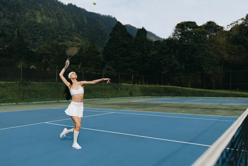 Young female tennis player serving on a blue court, wearing a sporty outfit, showcasing determination and focus amid a lush green background