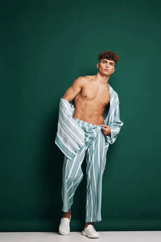 Young man with curly hair posing shirtless in striped pants and open jacket against green background. Fashion model showing confident expression and casual style in studio setting.