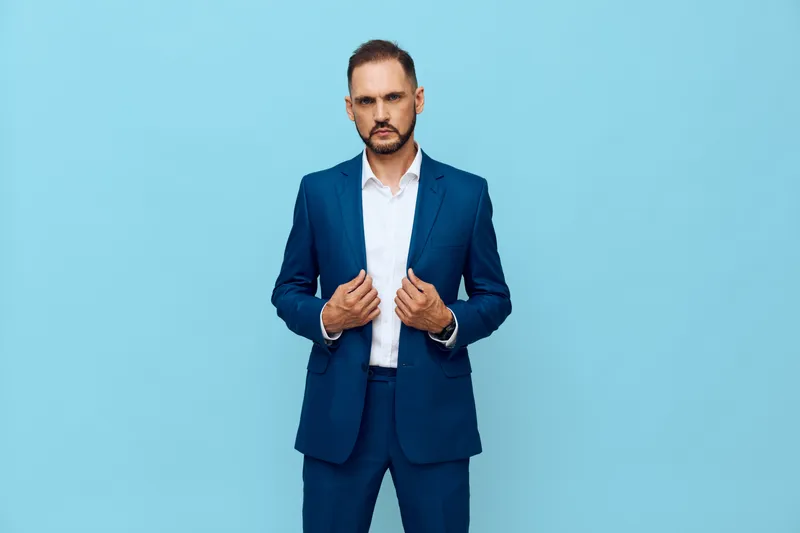 A professional man in a sharp blue suit stands against a solid blue backdrop, projecting confidence and calm as he adjusts his jacket with deliberate poise.