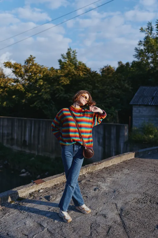 Young woman in her 20s wearing a colorful striped sweater and jeans with a crossbody bag, leaning and smiling outdoors by a rural fence in warm sunlight, casual travel lifestyle scene.