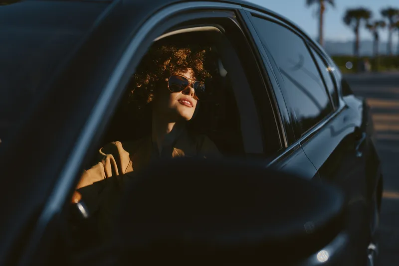 A stylish woman sits in a car with sunglasses, enjoying sunset light, embodying a relaxed lifestyle and a quiet late day drive in warm retro tones.