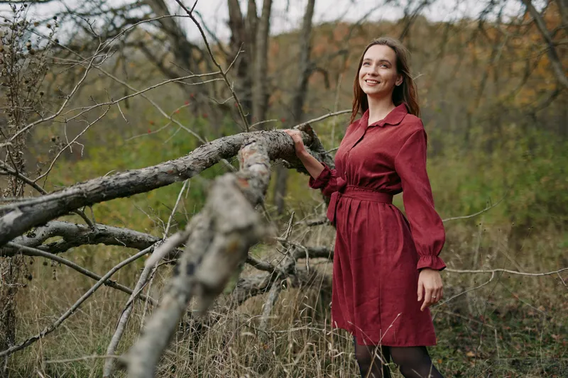 Portrait of a woman in a burgundy dress standing beside a fallen tree in a wild meadow, conveying authenticity and credibility through natural charm.
