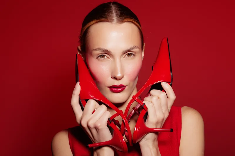 Portrait of a stylish woman holding red high heels near her face on red background. Fashion concept with bold makeup and elegant accessories in vibrant studio lighting.