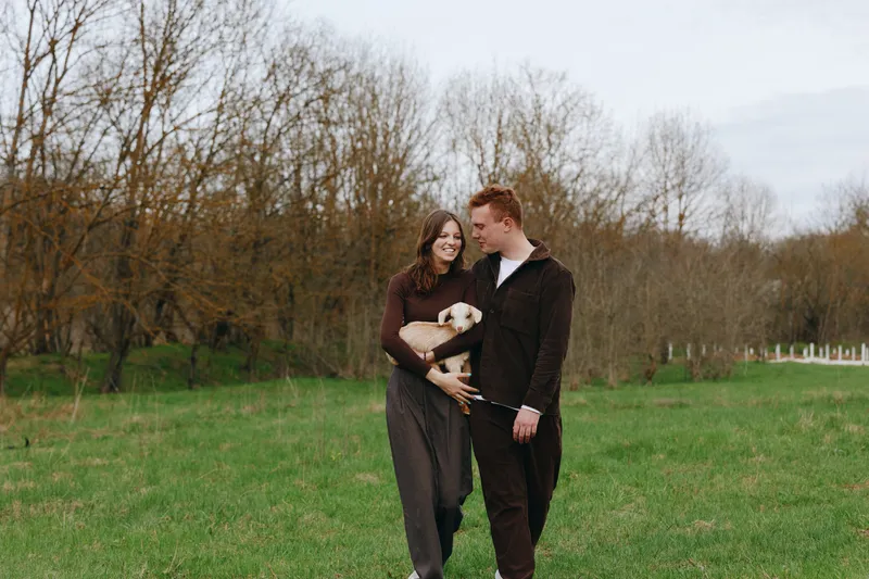 Happy couple strolling in a green park with a small dog, smiling and holding hands, enjoying a relaxed lifestyle moment, candid outdoor scene with trees in the background and soft natural light