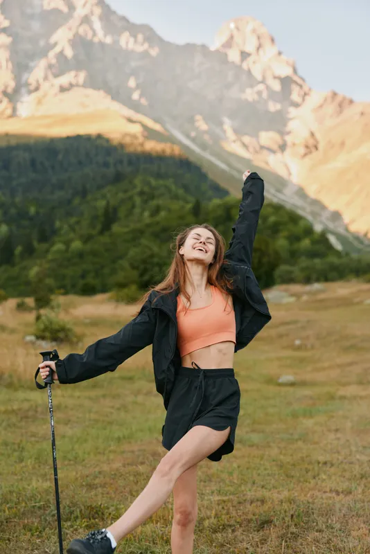 A cheerful young woman stands in a scenic mountain meadow, posing with a trekking pole. She wears a coral top and dark jacket, smiling with bright energy and confidence.