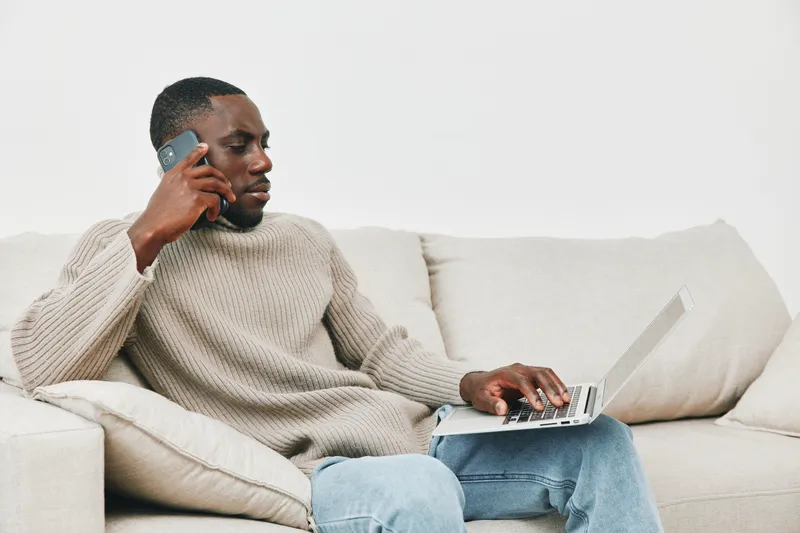 Young African American man working from home, wearing a beige sweater, engaged in a phone conversation while using a laptop on a cozy sofa