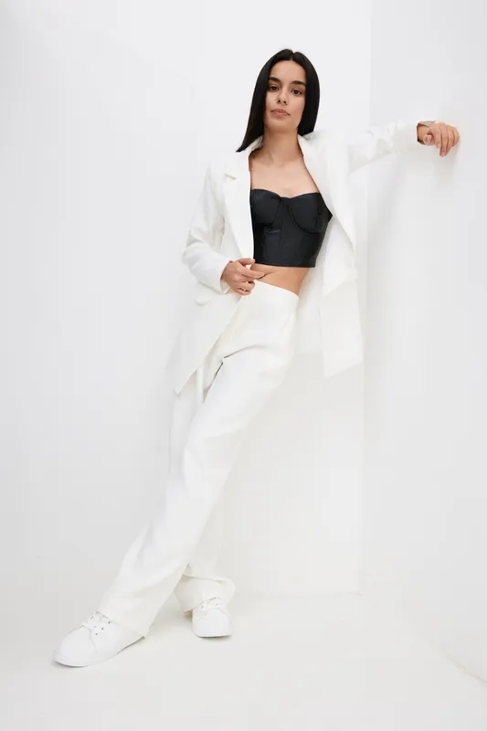 woman in white suit standing confidently against white wall wearing black crop top and white sneakers, poised, modern fashion style, minimalistic background