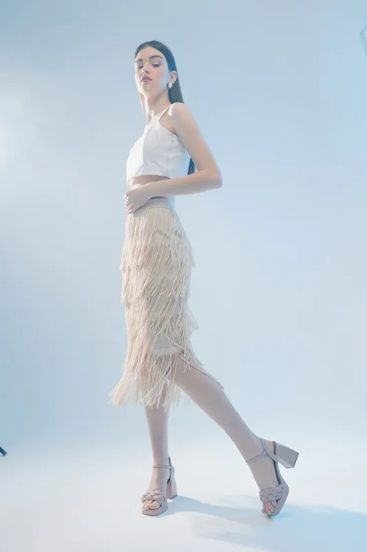 Young woman in a white top and beige fringed skirt poses confidently on a light background. Concept of woman beauty fashion and modern style in studio lighting.