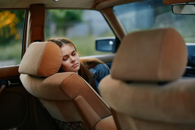 A young woman with long brown hair resting in a vintage car, showcasing a serene expression amidst a warm, sunny background.