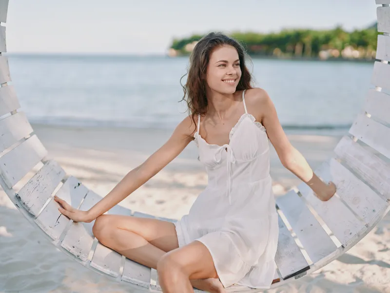 A smiling woman sits gracefully on a circular wooden swing by the sea, wearing a light white dress. Soft sunlight and a gentle breeze create a serene coastal mood with relaxed, carefree charm.