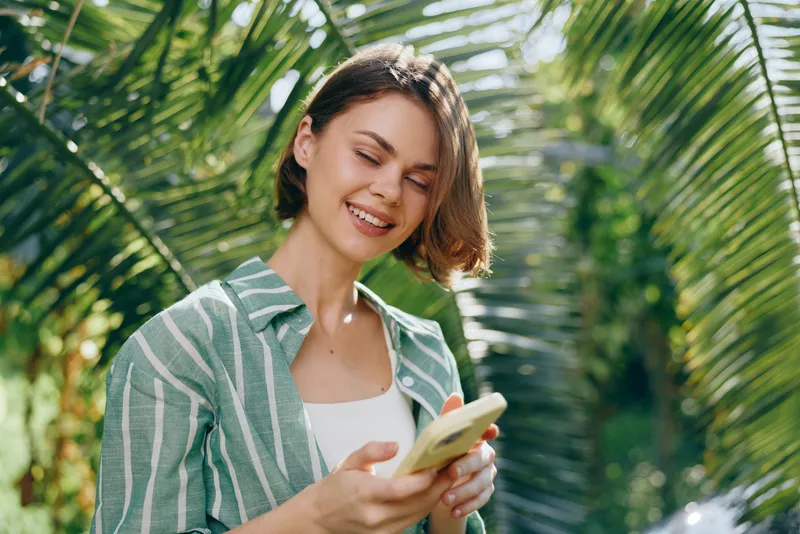 A joyful woman outdoors uses a smartphone among lush palm leaves and bright sunlight, capturing a candid moment.