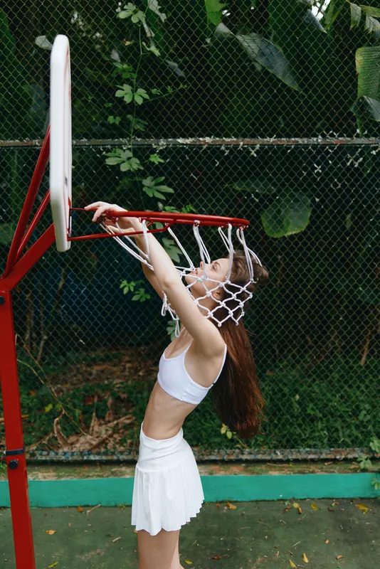Young athletic woman celebrating a basketball victory by hanging on the hoop, wearing a white sports outfit in a green outdoor court setting