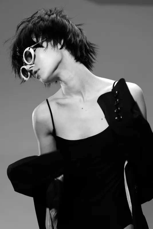 Black and white portrait of a stylish woman wearing sunglasses and a black dress with jacket, expressing a modern edgy vibe.
