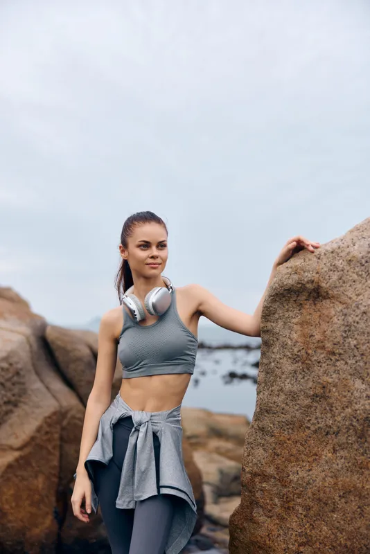Athletic young woman in sportswear standing by rocky beach, smiling confidently with headphones, conveying fitness and outdoor lifestyle