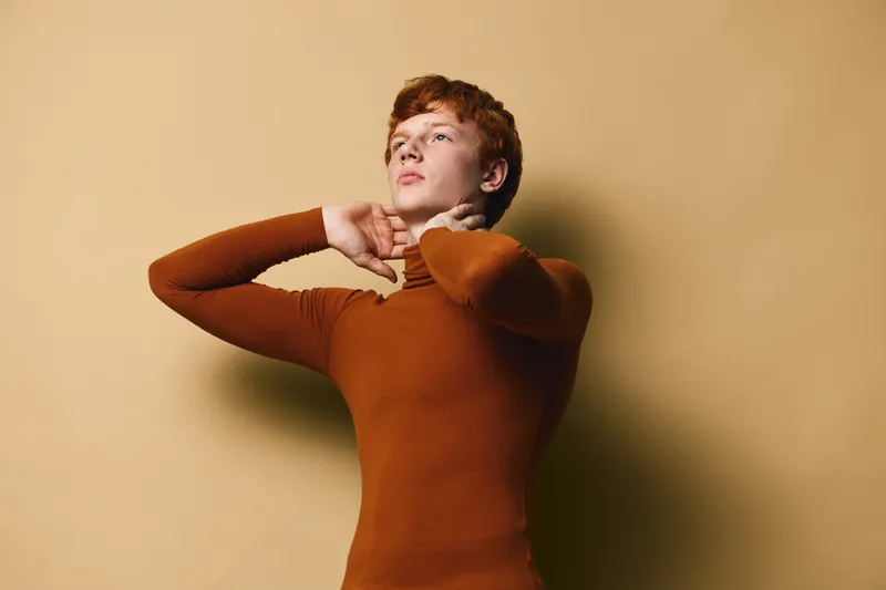 Young man with red hair wearing brown turtleneck posing with hands near neck against beige background. Stylish portrait, confident expression and warm colors in studio lighting.
