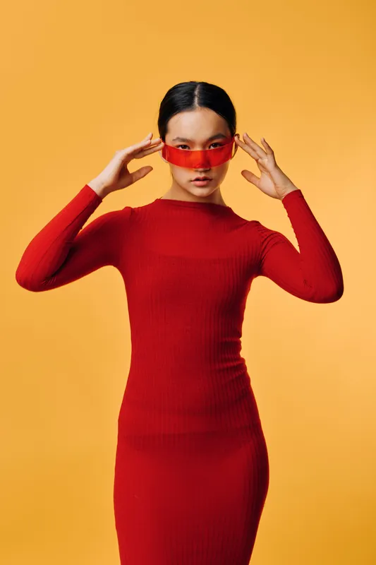 Woman in red dress and red sunglasses on orange background posing with hands near face. Fashion, style, portrait, trendy, confident model with sleek hair in studio lighting and modern outfit.