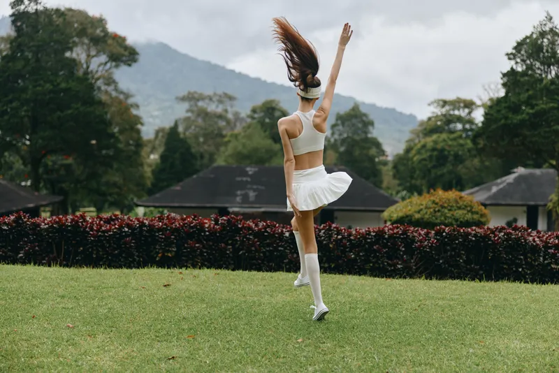 Energetic young woman in a white sporty outfit dancing joyfully in a green field, surrounded by trees and mountains, showcasing happiness and freedom