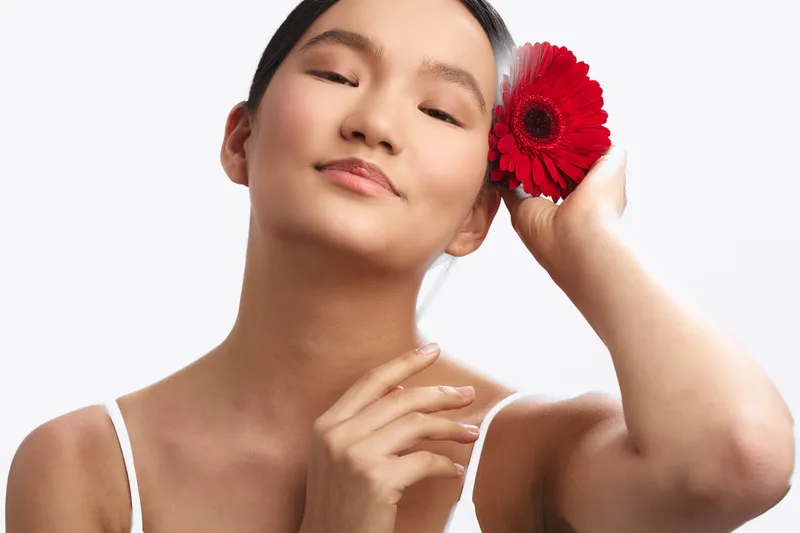 asian woman gently smiles while holding a red flower in a studio portrait, exuding grace and calm. soft lighting and serene mood create an elegant, beauty oriented atmosphere.