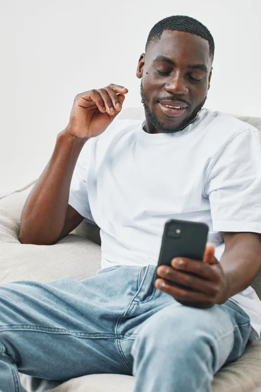 Young Black man smiling while using smartphone, wearing casual clothing in bright indoor setting, joyful expression shows enjoyment and modern lifestyle
