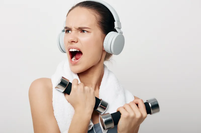 Young woman fitness portrait: athletic woman wearing headphones and a towel, holding two dumbbells and shouting with intense expression against a clean white background, sport concept.