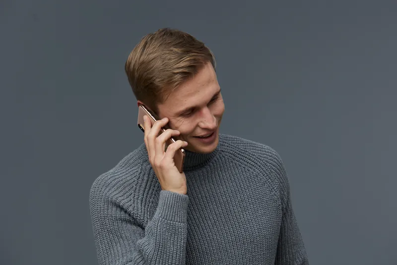 Smiling man talking on mobile phone, wearing casual gray sweater, looking down and smiling, isolated on plain gray background, studio portrait, lifestyle concept.