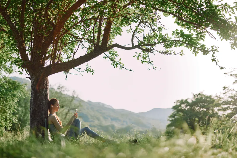 Young woman sitting under a tree, checking her phone in a peaceful outdoor setting, surrounded by greenery and sunlight, promoting relaxation and connection with nature