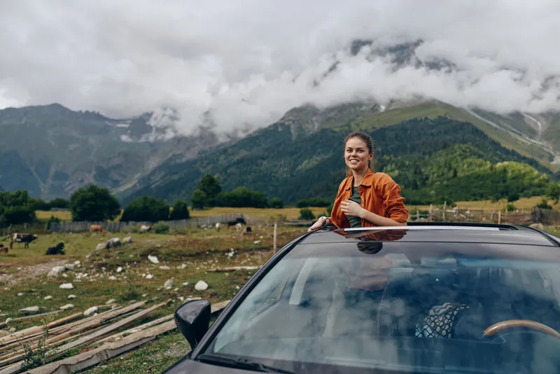 Woman in an orange jacket leaning out of a car sunroof in a scenic mountain landscape, smiling and enjoying the outdoors, capturing a carefree travel moment.