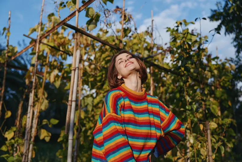 Woman in a rainbow sweater smiling in a garden by a wooden trellis, outdoor portrait of a young adult with short hair, relaxed pose, sunlit lifestyle and joyful mood.