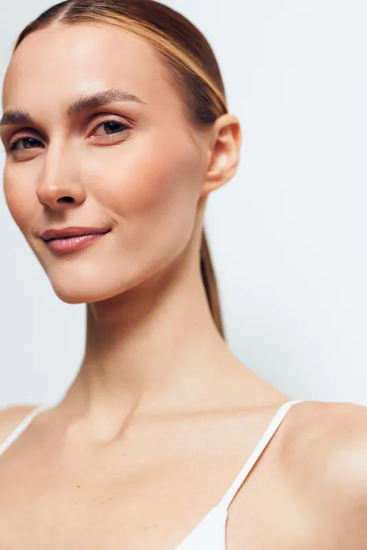 A confident young woman with clear skin and pulled-back hair smiles subtly against a plain white background. The portrait highlights natural beauty and healthy skin.
