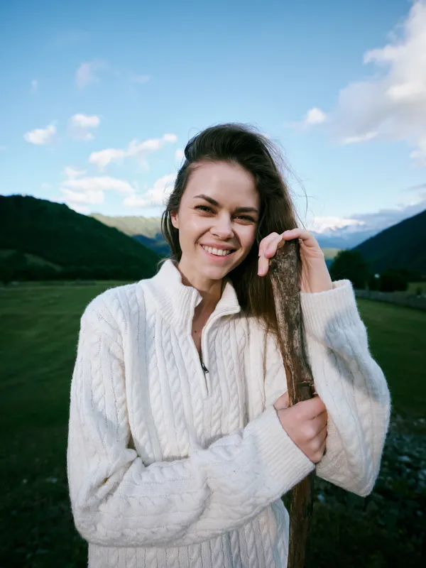 Smiling woman in a white knitted sweater stands in a valley with distant mountains, capturing a relaxed outdoor moment and natural charm under a blue sky.
