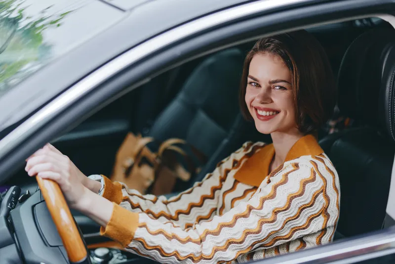 A cheerful woman driver sits behind the wheel, smiling warmly as she steers the car with confidence and focus, embodying safe, stylish travel.