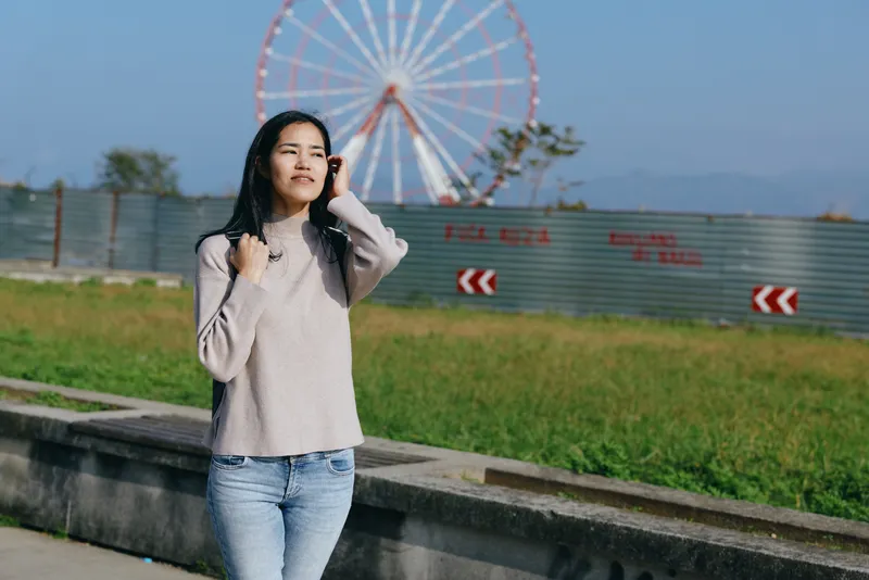 A woman talks on her phone outdoors near a ferris wheel in a park, wearing casual clothes under daylight, capturing a bright urban moment with greenery and open sky.