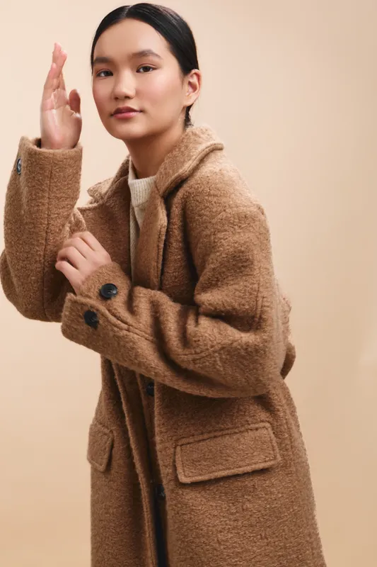 Asian woman wearing a cozy brown coat poses against a warm beige backdrop, exuding calm confidence and contemporary style in a fashion portrait.