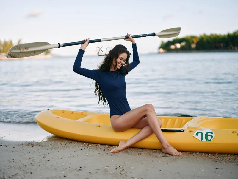 Young woman in a blue long sleeve swimsuit sits on a yellow kayak by the shore, lifting a paddle overhead with a joyful pose as calm water and distant trees frame the scene.