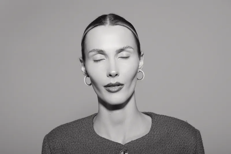 Young caucasian woman with pale skin and closed eyes poses calmly in studio, wearing hoop earrings and a knitted sweater.