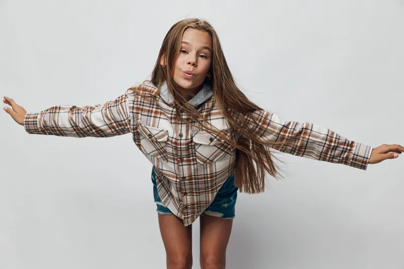 Girl in casual plaid shirt and denim shorts posing with arms outstretched and puckered lips against white background. Youthful model with long hair and playful expression in studio.