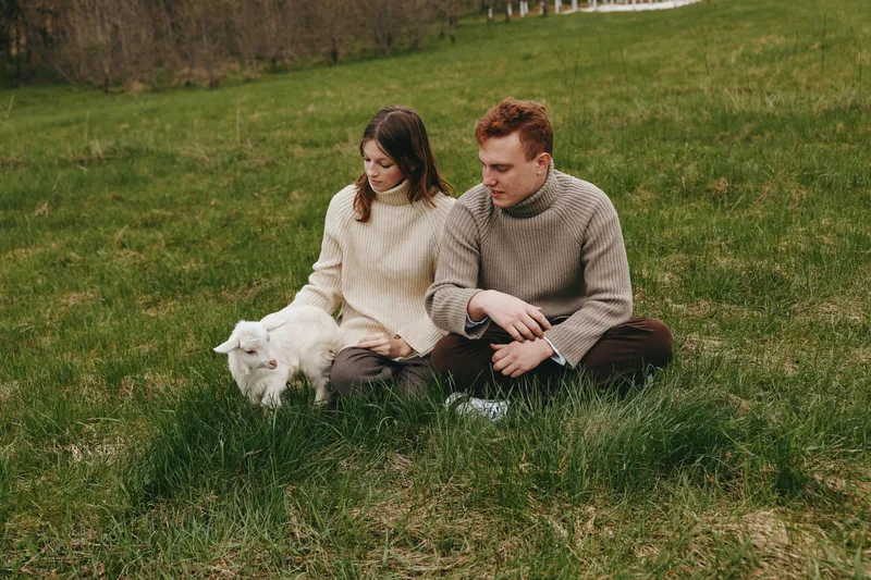 Couple outdoors on grass with a playful goat kid, cozy sweaters, soft light, calm countryside moment, relaxed posture, natural colors, shared affection