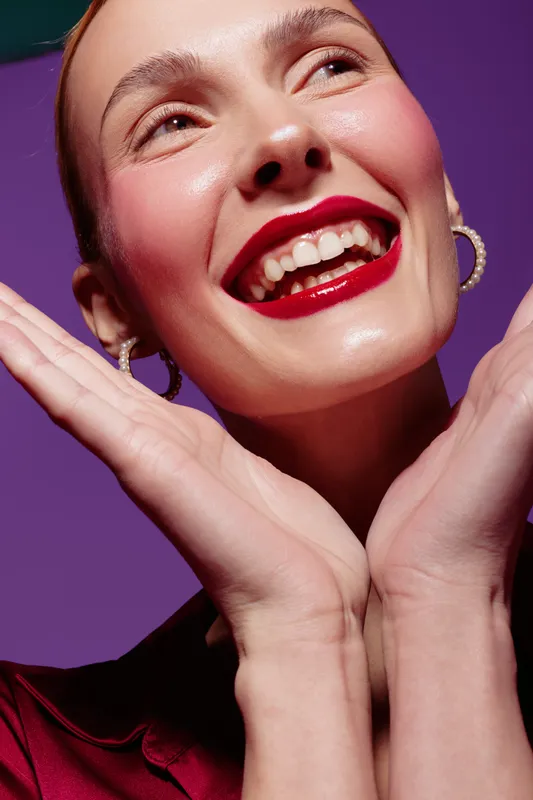 Close-up of happy woman with red lipstick smiling brightly, hands framing face. Studio portrait on purple background. Concept of beauty, makeup, joy, and positive emotions.