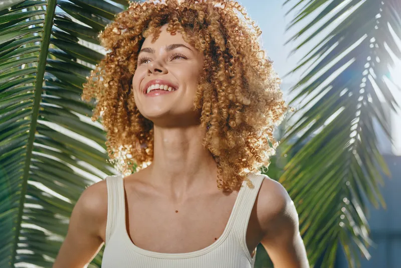 Smiling woman with curly hair enjoys sunny tropical background, wearing a white tank top, expressing happiness and natural beauty under palm leaves in bright daylight.