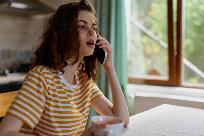 A young woman in a yellow striped t shirt sits at a sunlit kitchen table, talking on her phone with a thoughtful expression while holding a mug.