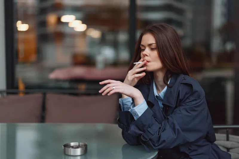 Young woman with long hair smoking cigarette outdoors at cafe table. Casual style with dark jacket, thoughtful expression, urban background with blurred lights.