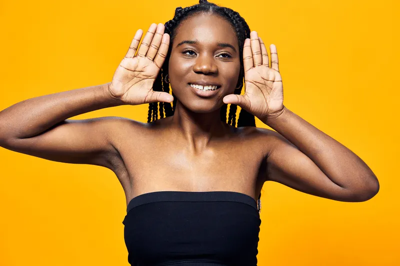 Smiling young Black woman with braided hair excitedly showing her hands against a bright yellow background, embodying joy and positivity