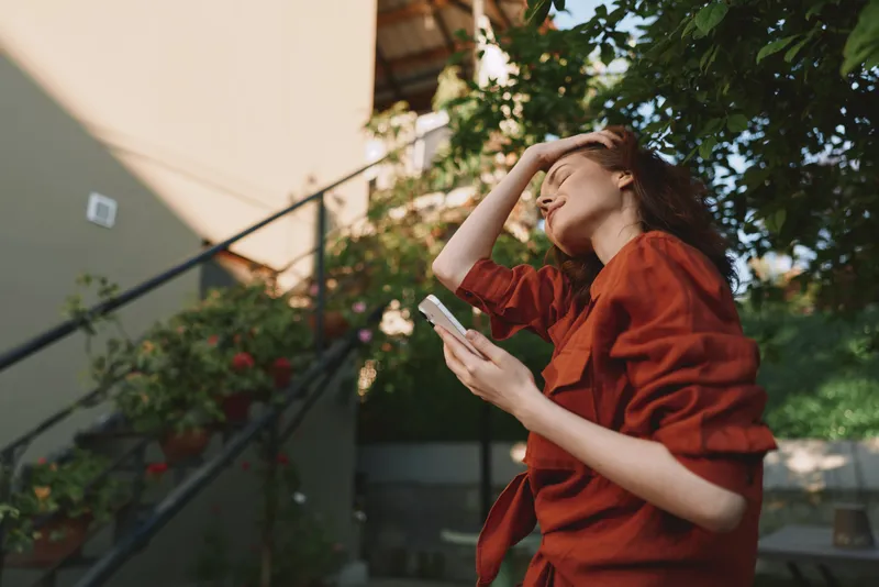 A woman in a red shirt stands outdoors, shielding her eyes from bright sun while scrolling on her phone, capturing a candid lifestyle moment on a warm day under clear skies