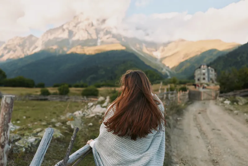 Woman with long hair wrapped in blanket looking at mountain landscape on a sunny day. Concept of lifestyle, travel, and nature exploration with soft natural light.