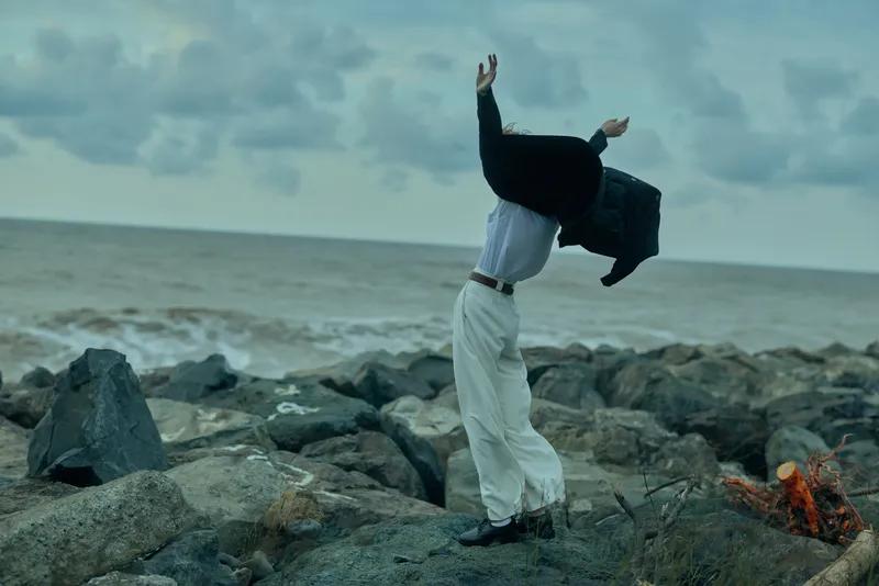 Windy coastal scene with a figure lifting a jacket over the head on a rocky shore, white pants and dark blazer, cloudy sky and choppy sea creating a dynamic, atmospheric composition.